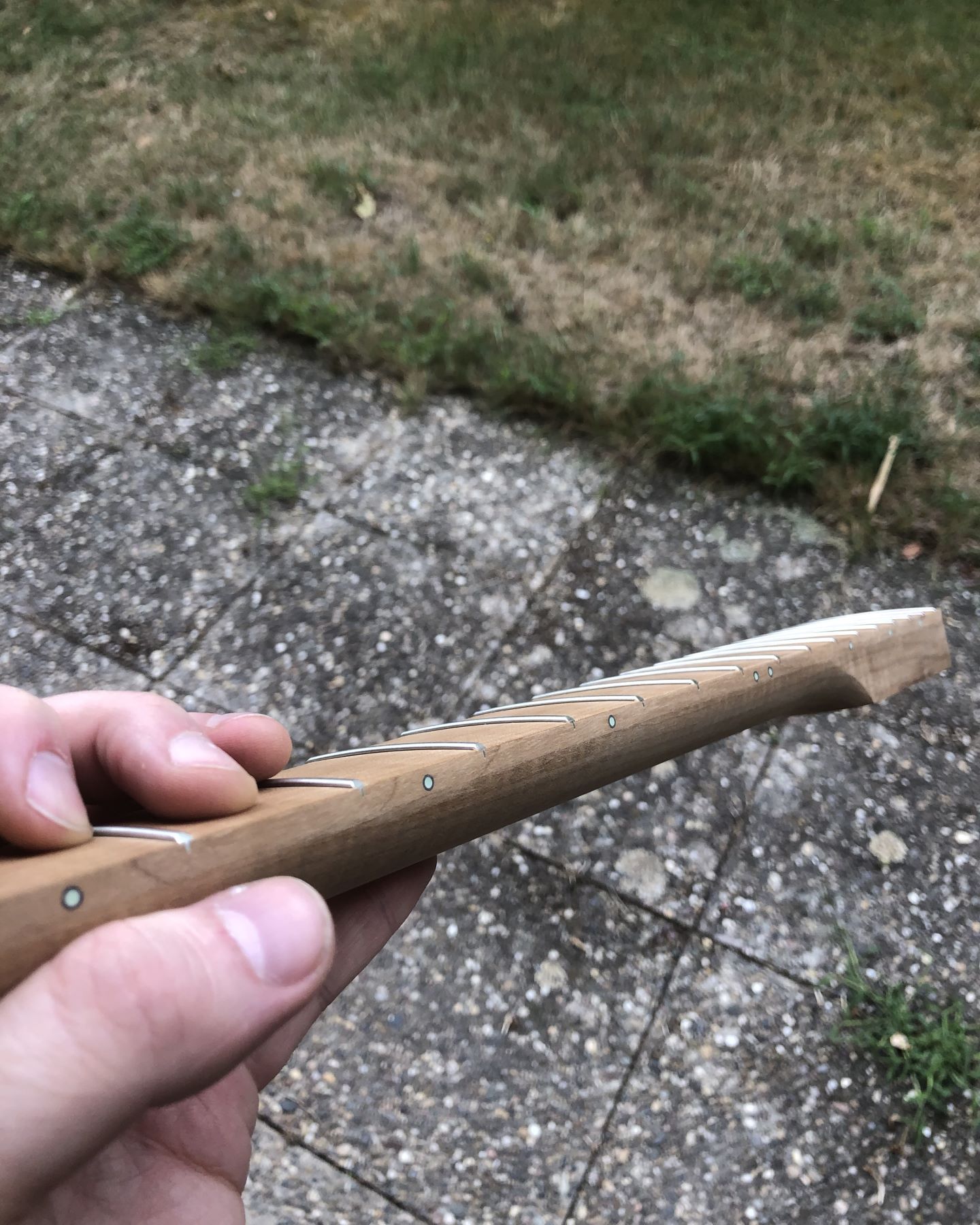 Photo showing the lower half of an unfinished roasted maple bass guitar neck not installed in a body with glow-in-the-dark side dot inlays with black rings around them for contrast.