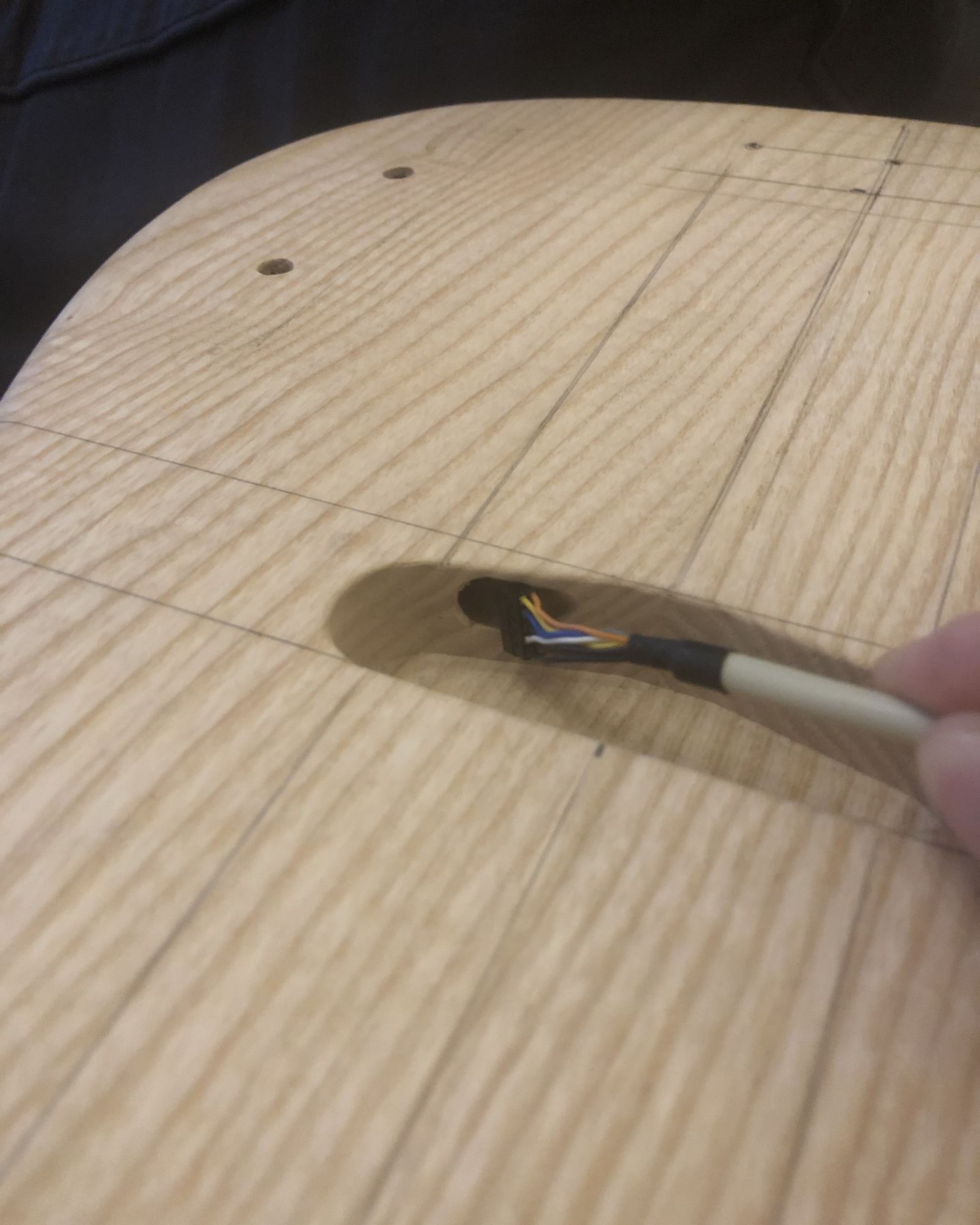 Photo of an unfinished ash bass guitar body with no pickup installed and a cable with a 10-pin 2 mm pitch connector held against the much smaller pre-drilled pickup wire hole.
