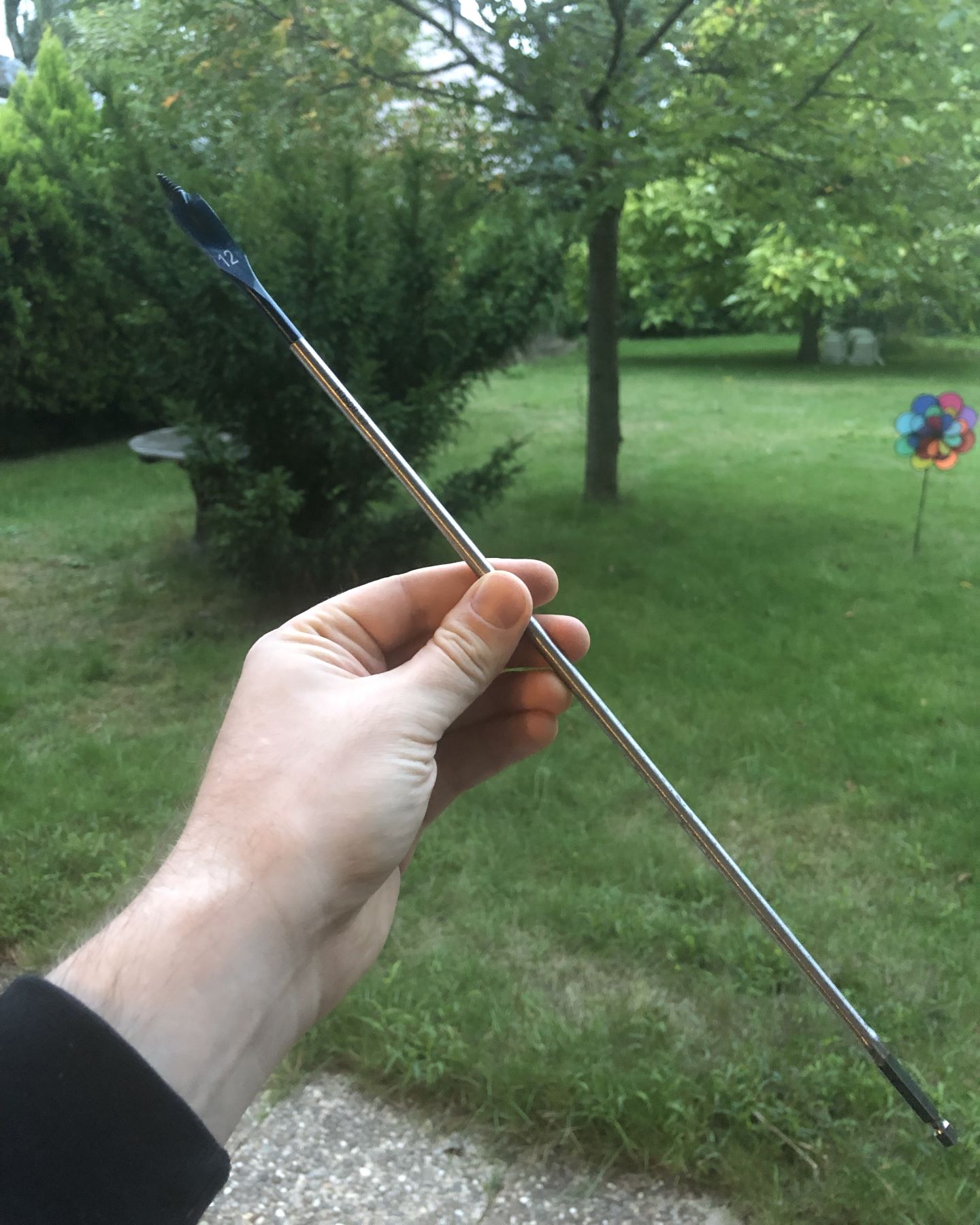 Photo of a 40 cm long, 12 mm spade drill bit, held up against the backdrop of a garden.