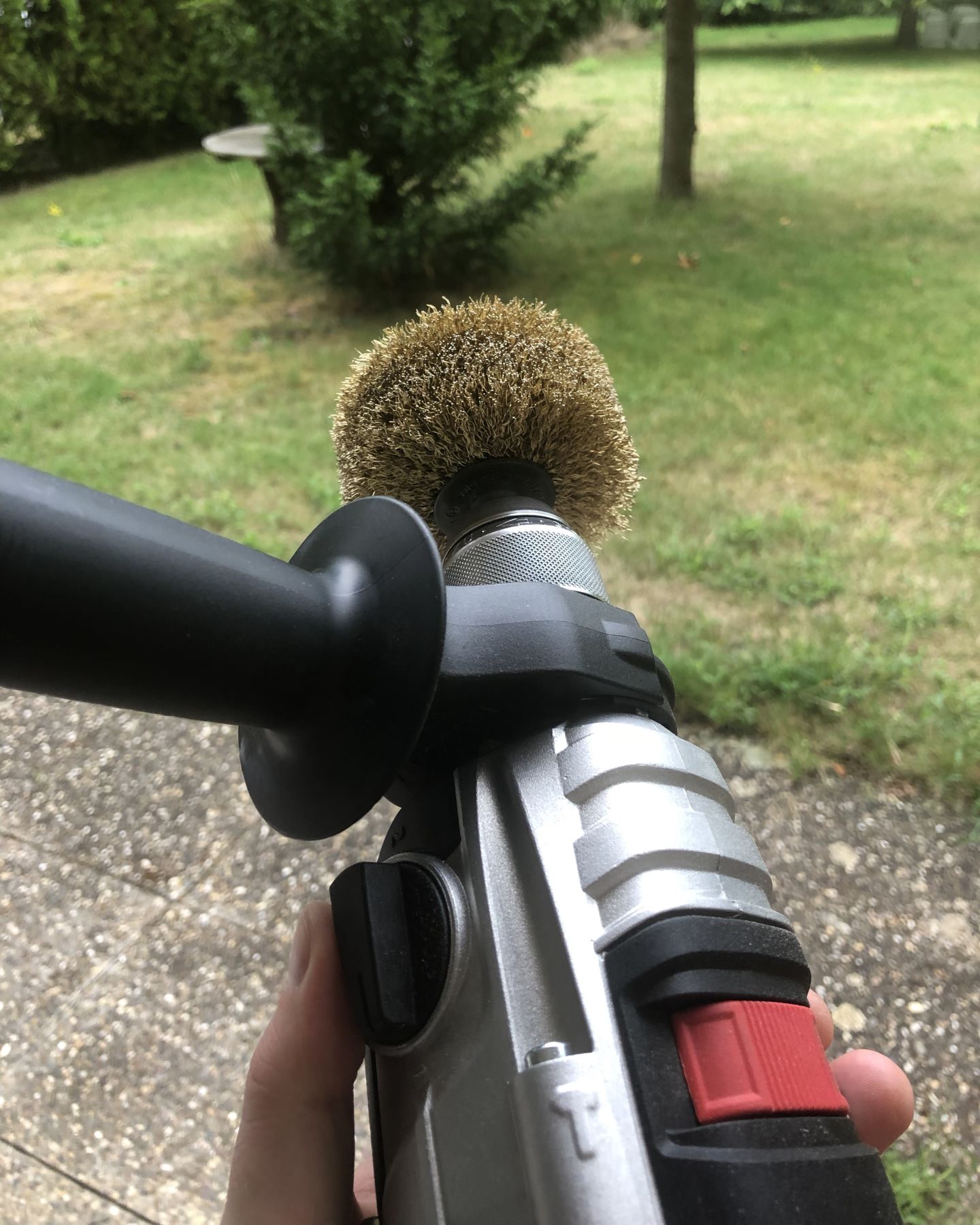 Photo of a large power drill with side handle installed and a large metal brush bit, held up against the backdrop of a garden.