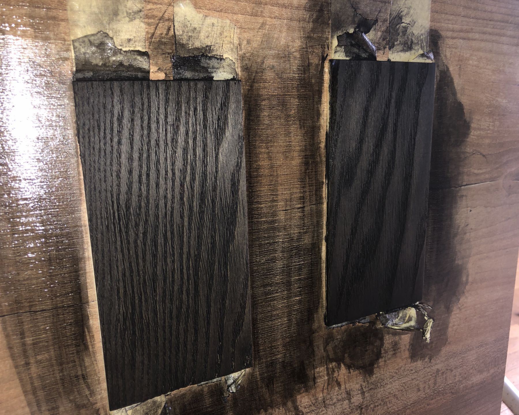 Photo of two flat pieces of ash stuck to another piece of wood with tape. The ash has pronounced wood grain and is stained black and covered in a semi-matte finish.