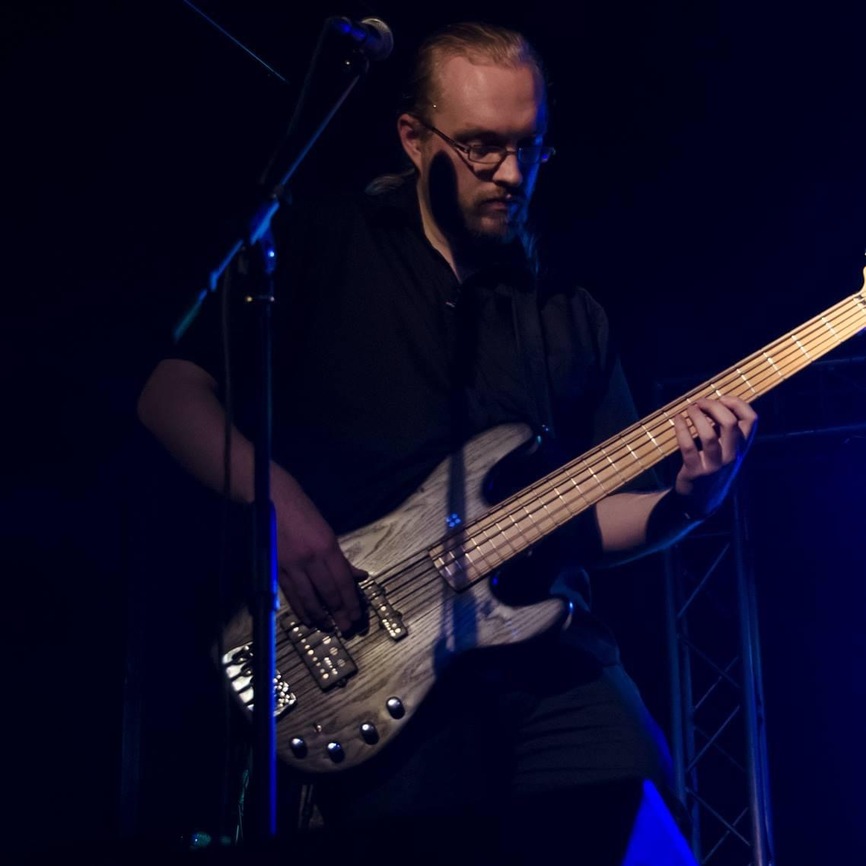 Photo of a long-haired blonde man with beard and glasses playing a black 5-string Sandberg bass on stage
